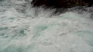 Fırtınalı deniz ve sahil kayalıklarının çok yakın görüntüsü etkileyici. Dalgalar taşlara güçlü bir şekilde çarparak beyaz köpük ve sprey püskürtür. Deniz suyu kayanın üzerine yuvarlanır ve geri döner. Yavaş çekim.