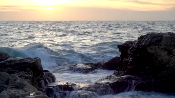 日没時の穏やかな海の景色 波は前景の海岸の岩の上を転がり 美しくそれらに対して破壊し スプレーの列を上げます 海の表面は地平線に進み オレンジ色の黄金の空と夕日の明るい光 — ストック動画