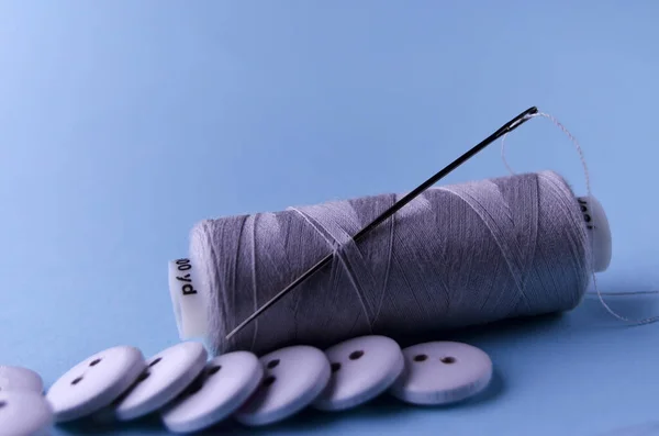 The needle is stuck in the bobbin of gray threads in the foreground scattered in a row of white wooden buttons