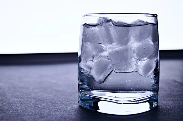 A glass full of carbonated water with ice cubes stands on a grey table against a background of counter light from an LED light close-up with a copy space