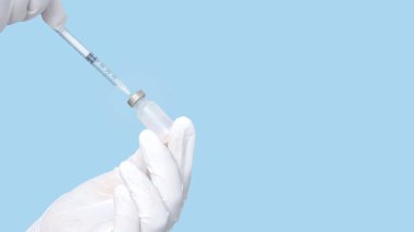 Aşı şişesi ve şırınga elinde mavi arka planda beyaz lastik eldivenler giyiyor..