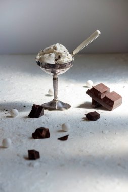 Gümüş bardakta tatlı, kaşık ve çikolata parçalı dondurma. Sert doğal ışıklandırmalı minimalist besin bileşimi.