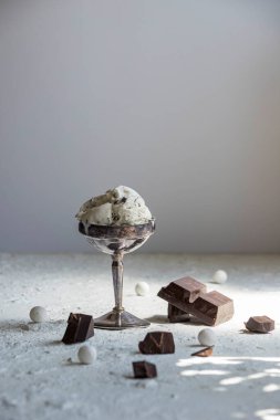 Gümüş bardakta vanilyalı dondurma ve dağınık siyah çikolatalı estetik natürmort. Sert beyaz yüzeyde sert güneş ışığı gölgeleri.