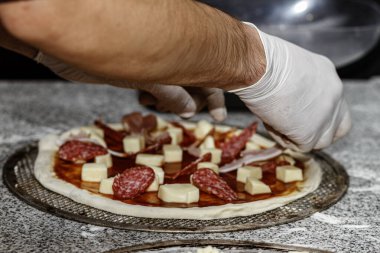 Şef eldivenlerle domates soslu ve peynirli çiğ pizzanın üzerine salam dilimleri düzenliyor. Et tepeleme süreci.