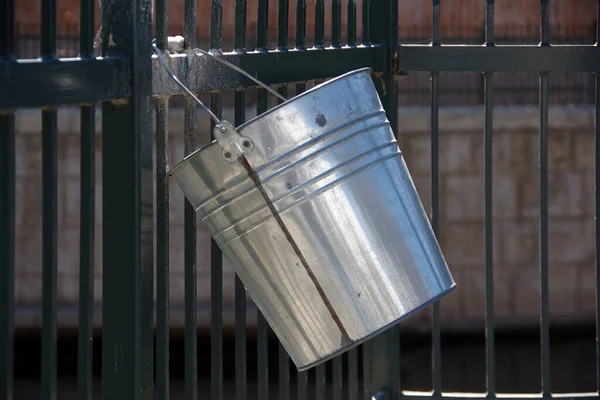 A silver colored metal bucket hanging on a fence over a channelled urban river