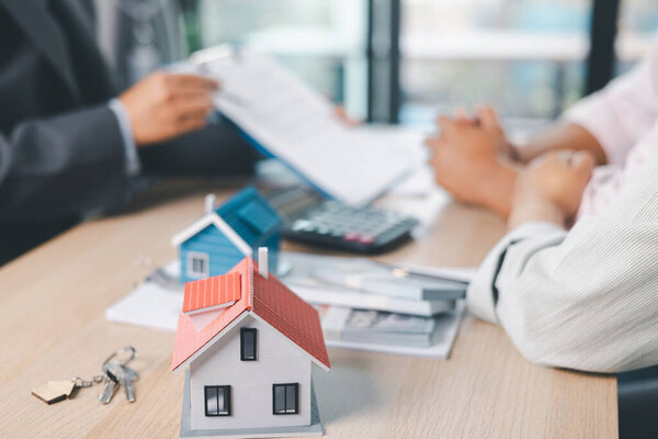 House model on desk with blurred people discussing contract in background, concept of real estate investment, mortgage loan, home ownership, property business and financial agreement.