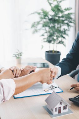 Emlakçı ve müşterinin el sıkıştığı ev modelleri, sözleşme belgeleri ve masa başındaki anahtarlar, başarılı emlak anlaşması, mortgage kredi anlaşması ve iş ortaklığı kavramları..