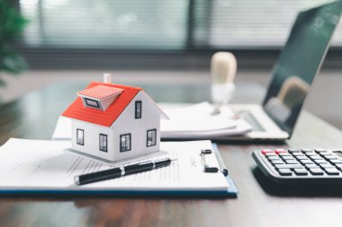 Masanın yanına yerleştirilen ev modeli madeni paraların yanı sıra, evrak işleri de gayrimenkul yatırımını, emlak değerini, mali planlama, mortgage seçeneklerini, emlak piyasasındaki pazar analizini simgeler..