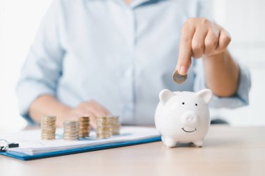 Tasarruf ve yatırım planlama kavramı. Bir iş kadını, mali güvenliği, bütçe yönetimini ve gelecekteki zenginliği sembolize eden belgelerle birlikte ofis masasındaki beyaz domuz kumbarasına para atıyor..