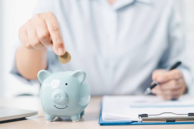 Tasarruf ve mali planlama kavramı. Bir iş kadını, yatırım, bütçe yönetimi ve gelecekteki büyümeyi sembolize eden belgeler ve dizüstü bilgisayarla birlikte ofis masasındaki mavi kumbaraya para koyar..