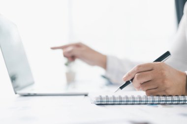 İş planlama ve finansal analiz kavramı. Bir iş kadını, modern ofis masasındaki bütçe yönetimi için dizüstü bilgisayar ve hesap makinesi kullanarak dosya ve raporlarla ilgili notlar yazar..