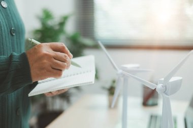 Güneş paneli ve rüzgar türbini modelinin yanında çalışan kişinin bulunduğu çevre dostu ofis sahnesi, yeşil işletmeleri, yenilenebilir enerji yeniliklerini ve sürdürülebilir teknoloji çalışma alanını temsil ediyor.