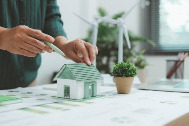 Masasında güneş paneli ve iş haritaları bulunan çevre dostu konut modeli, yeşil bina, yenilenebilir enerji, sürdürülebilir mimari ve çevresel yenilik kavramını sembolize ediyor.