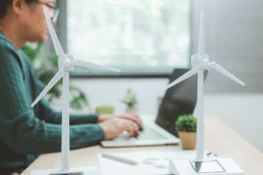 Güneş paneli ve rüzgar türbini modelinin yanında çalışan kişinin bulunduğu çevre dostu ofis sahnesi, yeşil işletmeleri, yenilenebilir enerji yeniliklerini ve sürdürülebilir teknoloji çalışma alanını temsil ediyor.