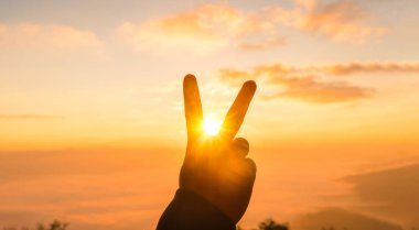 Silüet el barış işareti yapıyor. Güneşin altın gündoğumunda çerçeveleniyor. Sıcak turuncu gökyüzü ve yumuşak bulutlar. Zafer, özgürlük ve ilham için kopya alanı ile en az seyahat arkaplanı.