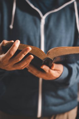 Elinde açık bir İncil tutan ve sayfaları çeviren adam, sıcak kapalı alan ışığı ve sığ alan derinliği, Hıristiyan inancını, bağlılığı, kutsal kitap çalışmasını ve evdeki sessiz ibadeti sembolize ediyor..