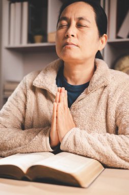 Asyalı bir kadın kapalı gözlerle ve ellerle açık bir kitabın üzerinde dua ediyor, sıcak bir iç mekan ışığı ve yumuşak bir odak, inanç, bağlılık, farkındalık ve evdeki huzurlu yansımayı ifade ediyor..