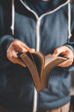 Elinde açık bir İncil tutan ve sayfaları çeviren adam, sıcak kapalı alan ışığı ve sığ alan derinliği, Hıristiyan inancını, bağlılığı, kutsal kitap çalışmasını ve evdeki sessiz ibadeti sembolize ediyor..