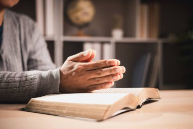 Açık bir kitap, sıcak bir ışık ve sığ bir alan derinliği üzerinde dua ederken, evde sessiz bir çalışma sırasında inanç, bağlılık, yansıma ve ruhsal rehberlik umudunu sembolize eder..