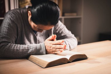 Asyalı bir kadın kapalı gözlerle ve ellerle açık bir kitabın üzerinde dua ediyor, sıcak bir iç mekan ışığı ve yumuşak bir odak, inanç, bağlılık, farkındalık ve evdeki huzurlu yansımayı ifade ediyor..