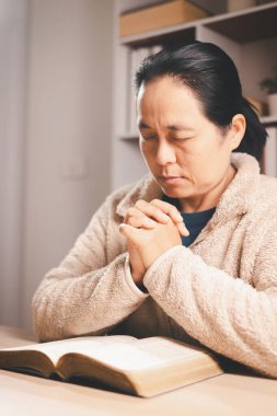 Asyalı bir kadın kapalı gözlerle ve ellerle açık bir kitabın üzerinde dua ediyor, sıcak bir iç mekan ışığı ve yumuşak bir odak, inanç, bağlılık, farkındalık ve evdeki huzurlu yansımayı ifade ediyor..