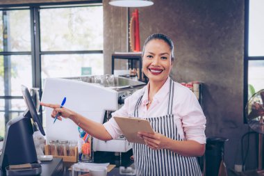  Önlüklü kafe sahibi sipariş verdi. Tezgahın arkasında duran kendine güvenen kadın baristanın portresi. Önlüklü kadın kafe sahibi kameraya bakıyor ve gülümsüyor..