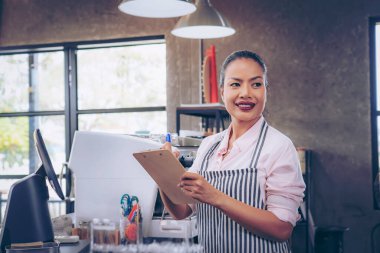  Önlüklü kafe sahibi sipariş verdi. Tezgahın arkasında duran kendine güvenen kadın baristanın portresi. Önlüklü kadın kafe sahibi kameraya bakıyor ve gülümsüyor..