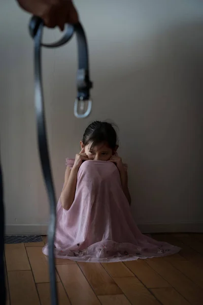 手にベルトが悲しみを脅かす少女を怖がらせた 光のドアの前の暗い廊下に座っている子供 恐怖と優柔不断だ 子供の概念に対する家庭内暴力 — ストック写真