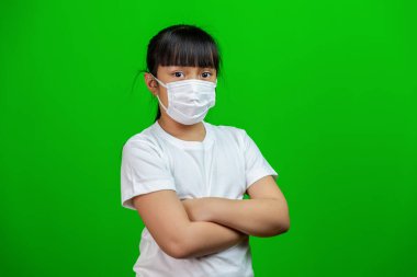 Koronavirüsü korumak için beyaz tişörtlü Asyalı kız çocuğu, Covid-19. Yeşil arka planda kollarını kavuşturuyor.                                                                                                                               