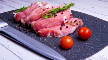 Domuz eti. Siyah kesim tahtasında taze domuz bifteği ve baharatlar, kiraz domatesleri, biberiye, biber, biber, bıçak, kopyalama alanı.