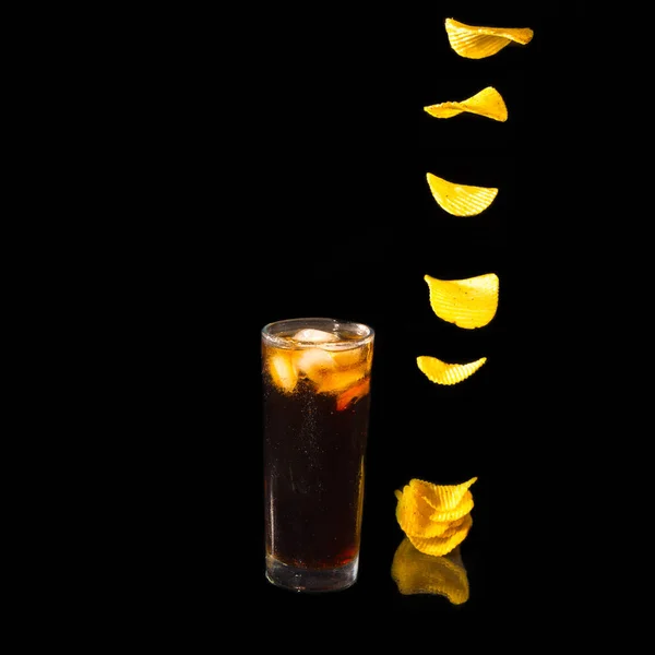 A glass of Pepsi Cola with ice and potato chips fall on the table in a stack on a black background, copy space, levitation