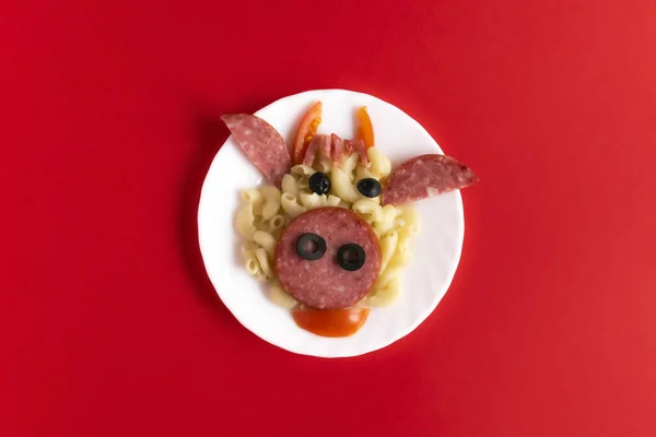 Lustiges Frühstück für Kinder in Form eines Stiergesichts aus Nudeln, Wurst, Speck, Oliven und frischen Tomaten auf rotem Hintergrund. Anleitung, wie man kreatives Food-Art-Frühstück für Kinder macht. — Stockfoto