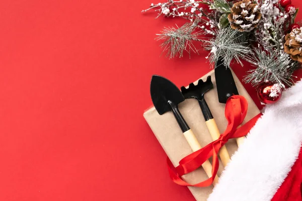 Noel ve yeni yıl kompozisyonu bahçe aletleri, kürek ve tırmık ve kırmızı arka planda Noel Baba şapkası. Bahçe işleri ve kart, takvim ve kapsama alanları için çiftçilik. Üst manzara, düz uzanma