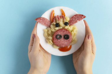 Çocuklar için komik kahvaltı, öğle yemeği ya da akşam yemeği makarna, sosis, pastırma, zeytin ve mavi arka planda taze domateslerden yapılmış boğa suratı şeklinde. Yaratıcı yemek sanatı kahvaltısı hazırlayan kadın elleri tabağı tutuyor. Üst görünüm.