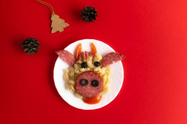 Çocuklar için makarna, sosis, domuz pastırması, zeytin ve kırmızı zemin üzerinde taze domateslerden yapılmış boğa suratı şeklinde komik bir kahvaltı. Koni ağacından Noel ağacı olan çocuklar için yemek sanatı tabağı. Üst görünüm.