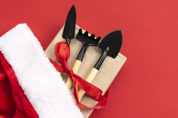 Noel ve yeni yıl kompozisyonu bahçe aletleri, kürek ve tırmık ve kırmızı arka planda Noel Baba şapkası. Bahçe işleri ve kart, takvim ve kapsama alanları için çiftçilik. Üst manzara, düz uzanma