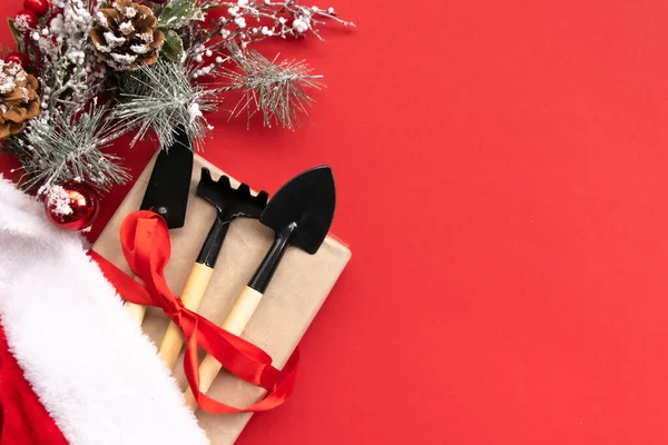 Noel ve yeni yıl kompozisyonu bahçe aletleri, kürek ve tırmık ve kırmızı arka planda Noel Baba şapkası. Bahçe işleri ve kart, takvim ve kapsama alanları için çiftçilik. Üst manzara, düz uzanma
