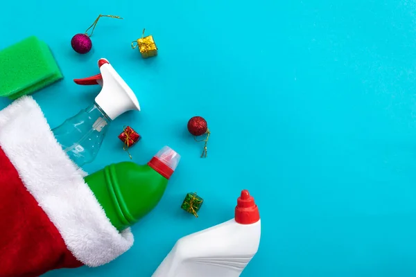 Noel hediyesi olan Noel Baba şapkası. Mavi arka planda ev temizleme malzemeleri. Temizleme sıvısı ve süngerli sıra sıra plastik şişeler, üstüne de kopyalama alanı. Temizlik işi, üretim, alışveriş ve teslimat.