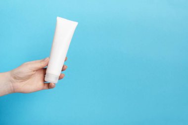 Kadınların elinde mavi arka planda beyaz krema tüpü var. Güzellik konsepti, temiz ve bakımlı kozmetik.