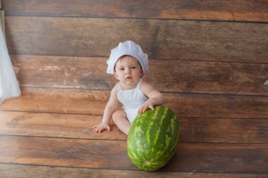 Bir yaşında bir kız elinde karpuzla oturuyor. Şef kostümü giymişti..