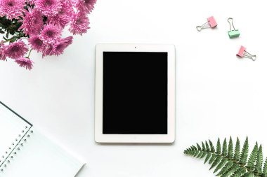 Tablet bilgisayar, defter, ataç, pembe kasımpatı buketi ve beyaz arka planda eğreltiotu yaprakları. Samimi iş yeri, üst görünüm, kopyalama alanı.