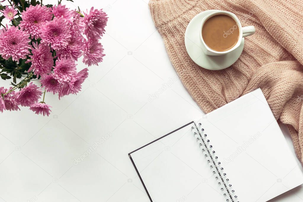 Cozy female workplace with a blank notebook, pen, cup of coffee, knitted sweater and a bouquet of chrysanthemum flowers. White background, top view, flat lay, copy space.