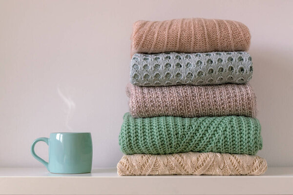 Stack of warm cozy knitted womens sweaters and cup of hot drink on white background. Autumn or winter comfort concept