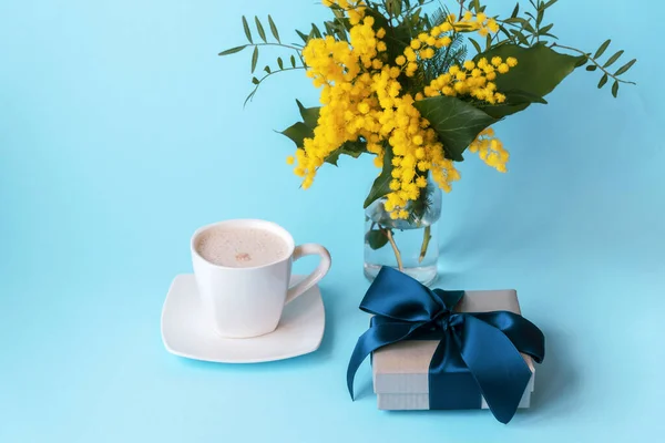 Cup of coffee, gift in a box and yellow spring flowers on a blue background. Valentines day or womens day concept.