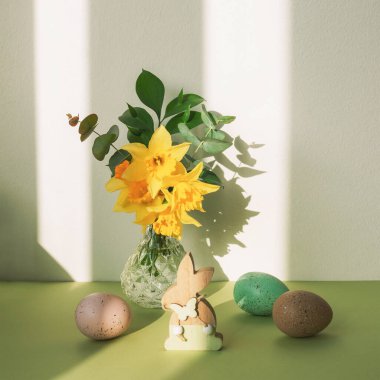 Tahta oyuncak tavşancık ve paskalya yumurtası, yeşil bir masanın üzerindeki bahar çiçekleri güneş ışığında beyaz bir duvara karşı.
