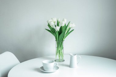 Cam vazoda bir buket beyaz lale, beyaz bir masada bir fincan ve bir sürahi süt. Sabah, kahvaltı, bahar konsepti.