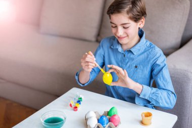 Dokuz yaşında mavi gömlekli bir kız, beyaz bir masada yumurta boyuyor. Paskalya konsepti. Seçici odak.