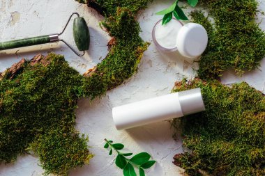 Nemlendirici krem kavanozu, serum ve masaj rulosu beyaz arka planda yosun ve yeşillikle birlikte. Doğal organik kozmetik kavramı. Üst manzara, düz uzanma.