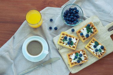 Kremalı ve yaban mersinli waffle, bir fincan kahve ve portakal suyu bez peçetenin üzerinde. Üst manzara, düz uzanma.