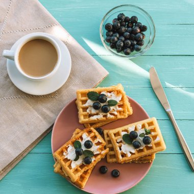Turkuaz masadaki pembe tabakta krem peynirli ve yaban mersinli bir fincan kahve ve waffle. Üst manzara, düz uzanma.